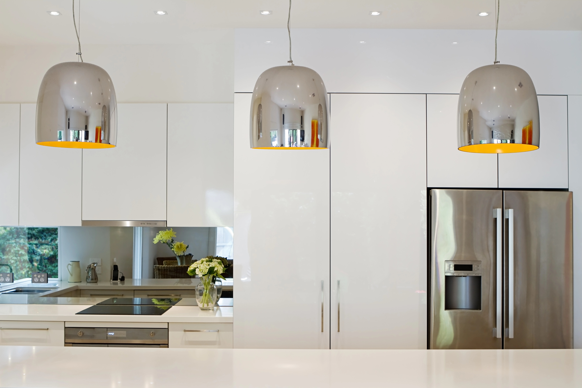 Modern kitchen with white cabinets, stainless steel fridge, built-in stove, and three metallic pendant lights that shine brilliantly after a recent electrical remodeling.