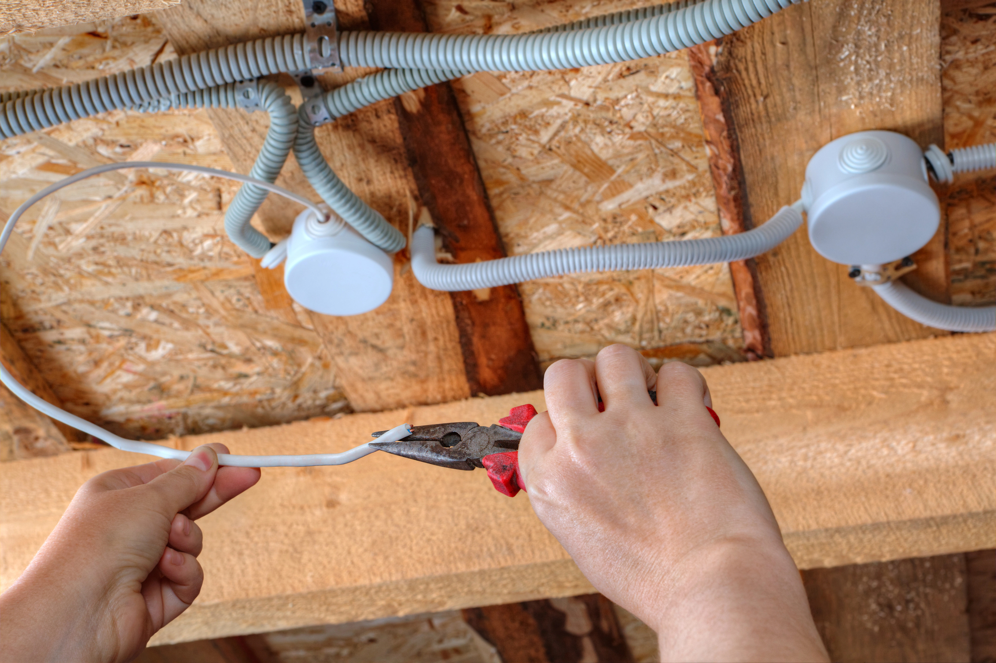 Hands using pliers to cut electrical wire near exposed ceiling fixtures on a wooden beam.