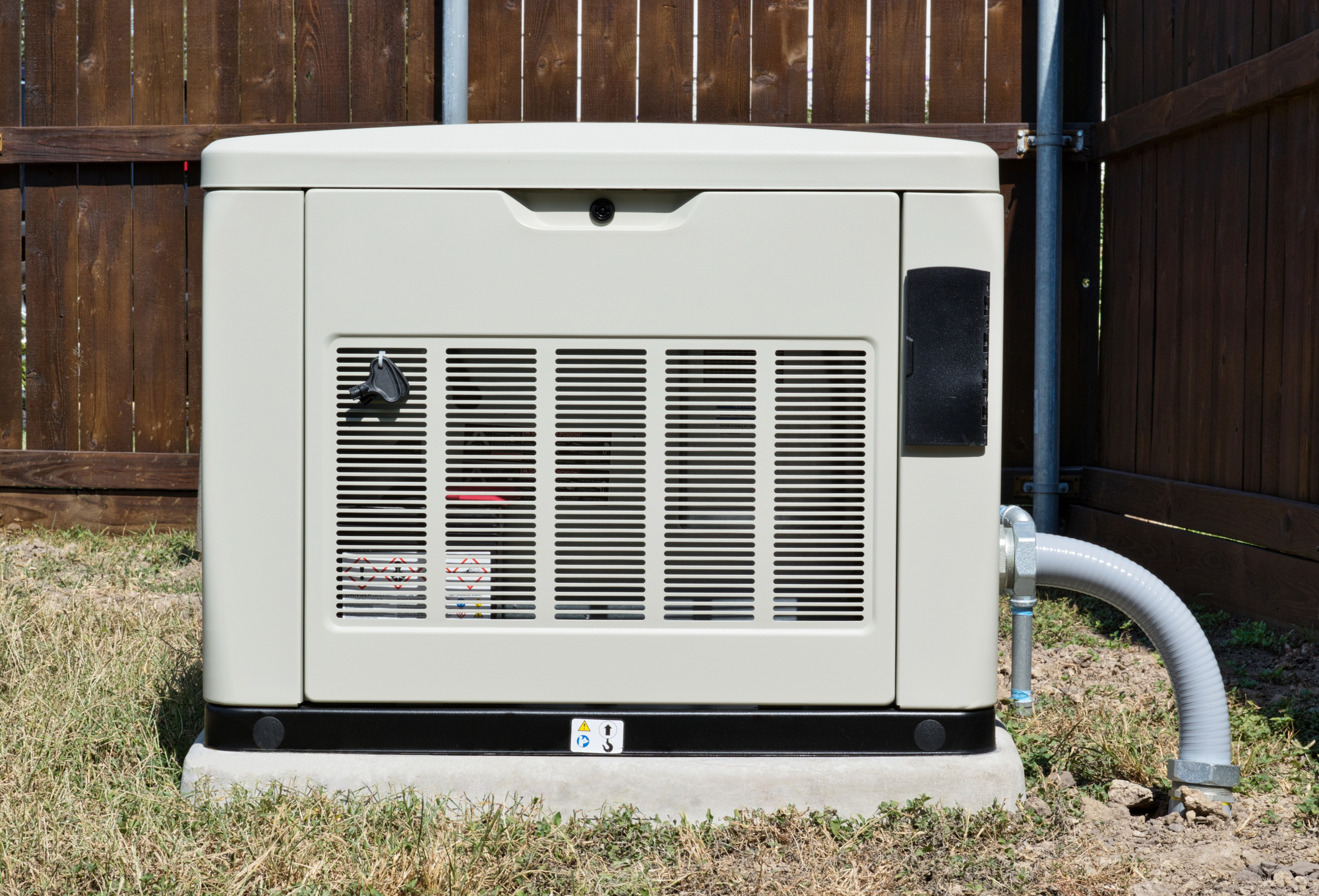 A beige outdoor generator sits on a concrete base in a grassy area, enclosed by a wooden fence.